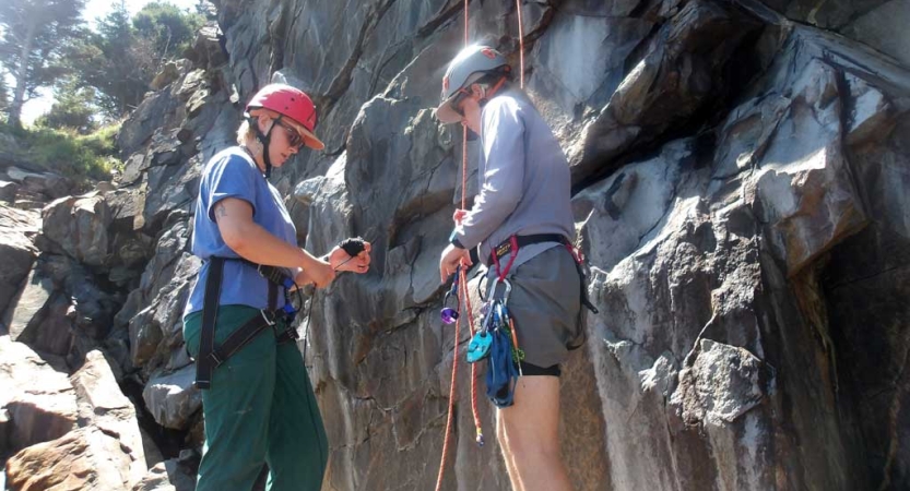 Two people wearing safety gear stand at the bottom of a rock wall, preparing to climb. 